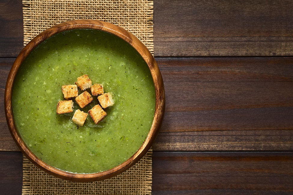 Fresh cream of zucchini soup with croutons in wooden bowl, photographed overhead on dark wood with natural light (Selective Focus, Focus on the soup)